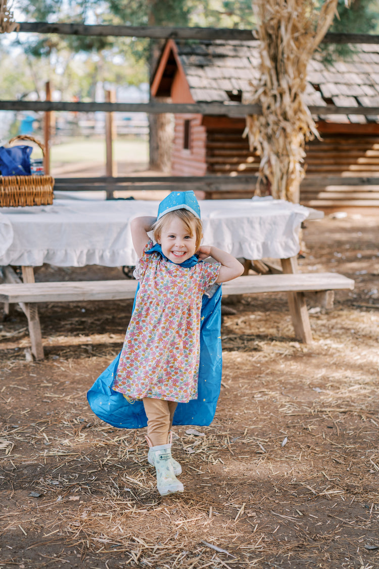 Child in blue cape walking in nature setting with table in the background. 