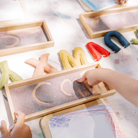 Children playing with wooden letter molds filled with colored sand on a light surface.