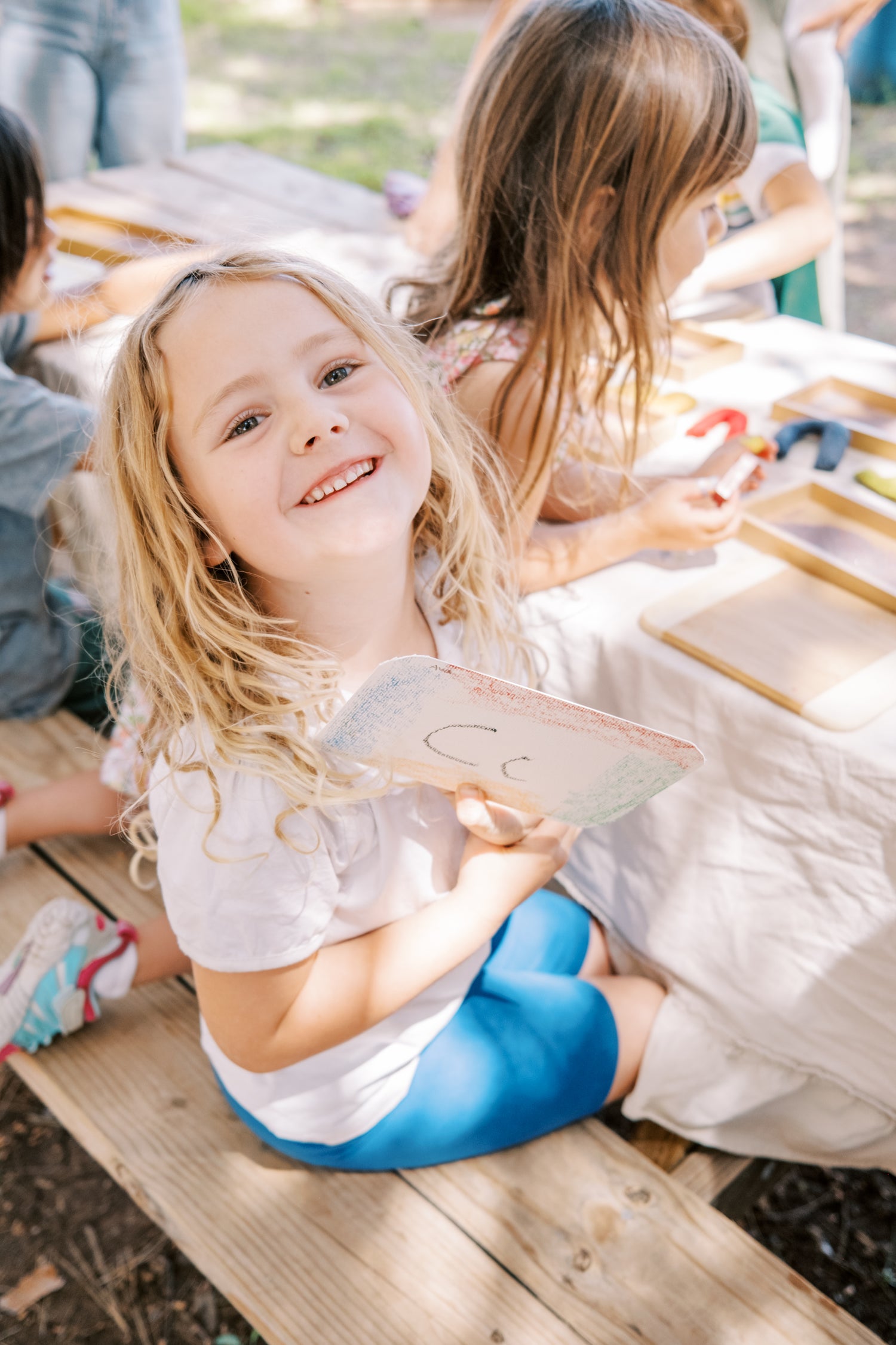 Young girl sitting at a wooden table outdoors, holding a small object.