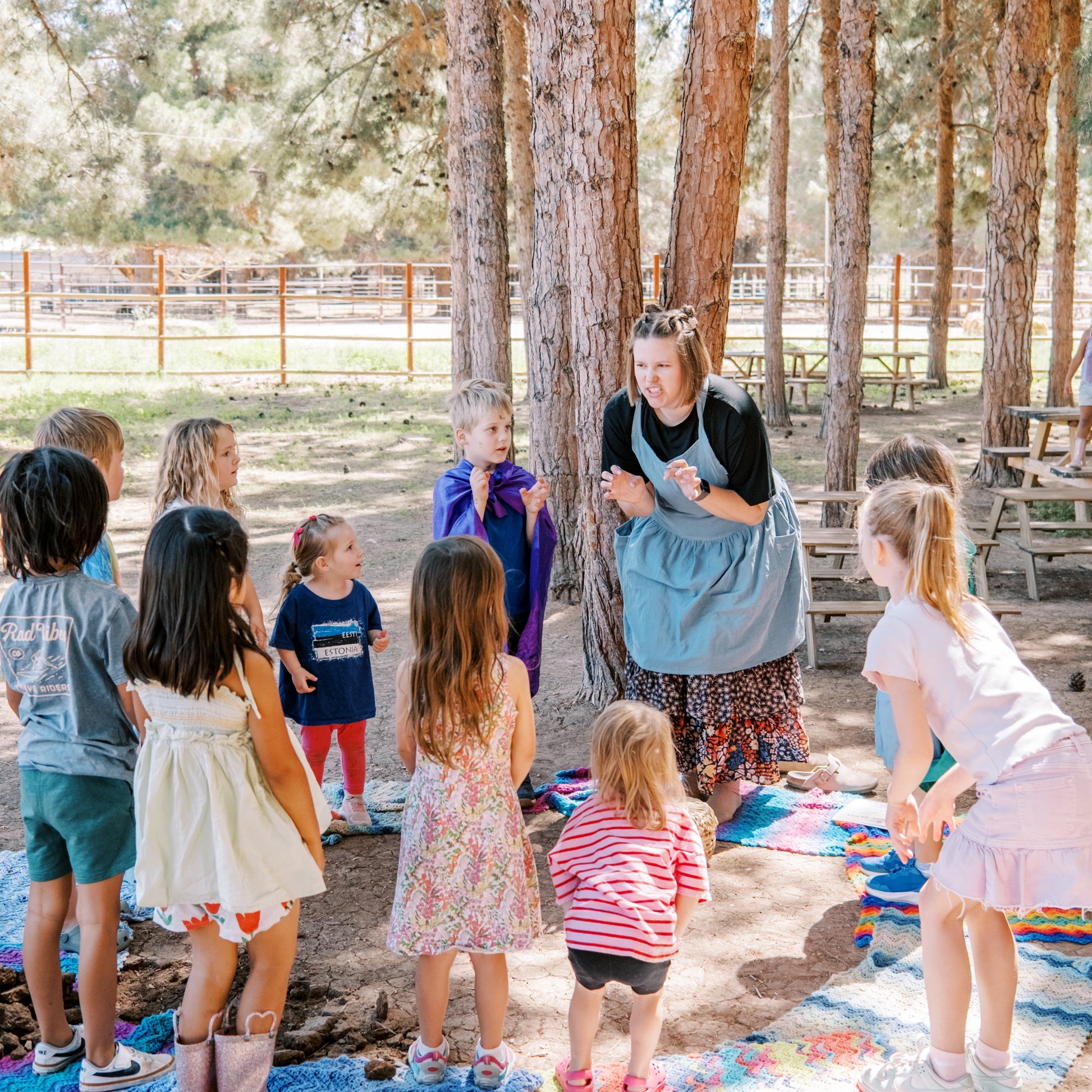 Children and a teacher gathered outdoors on a blanket with trees in the background