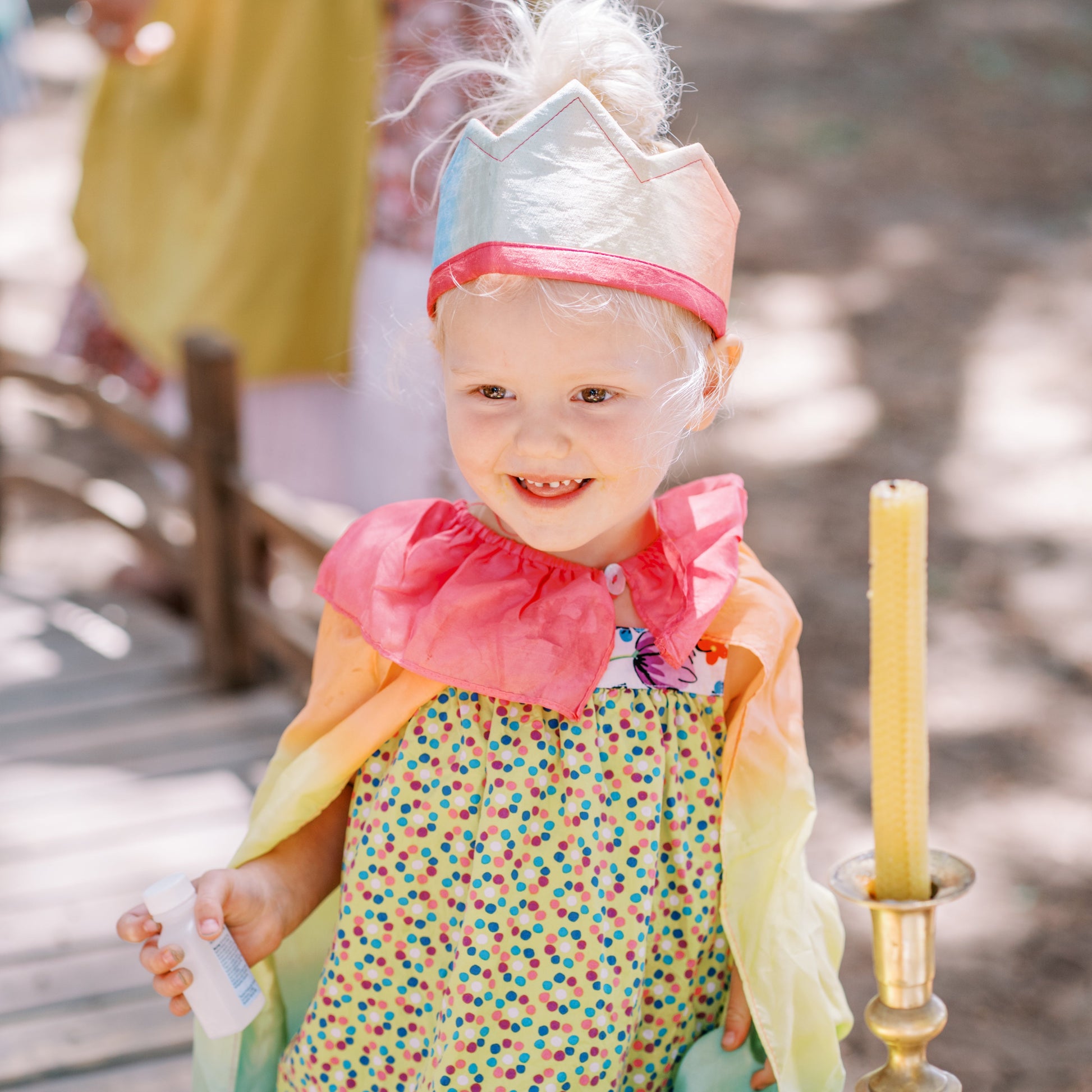 Child in colorful outfit with decorative headpiece near a candle and table setting outdoors.