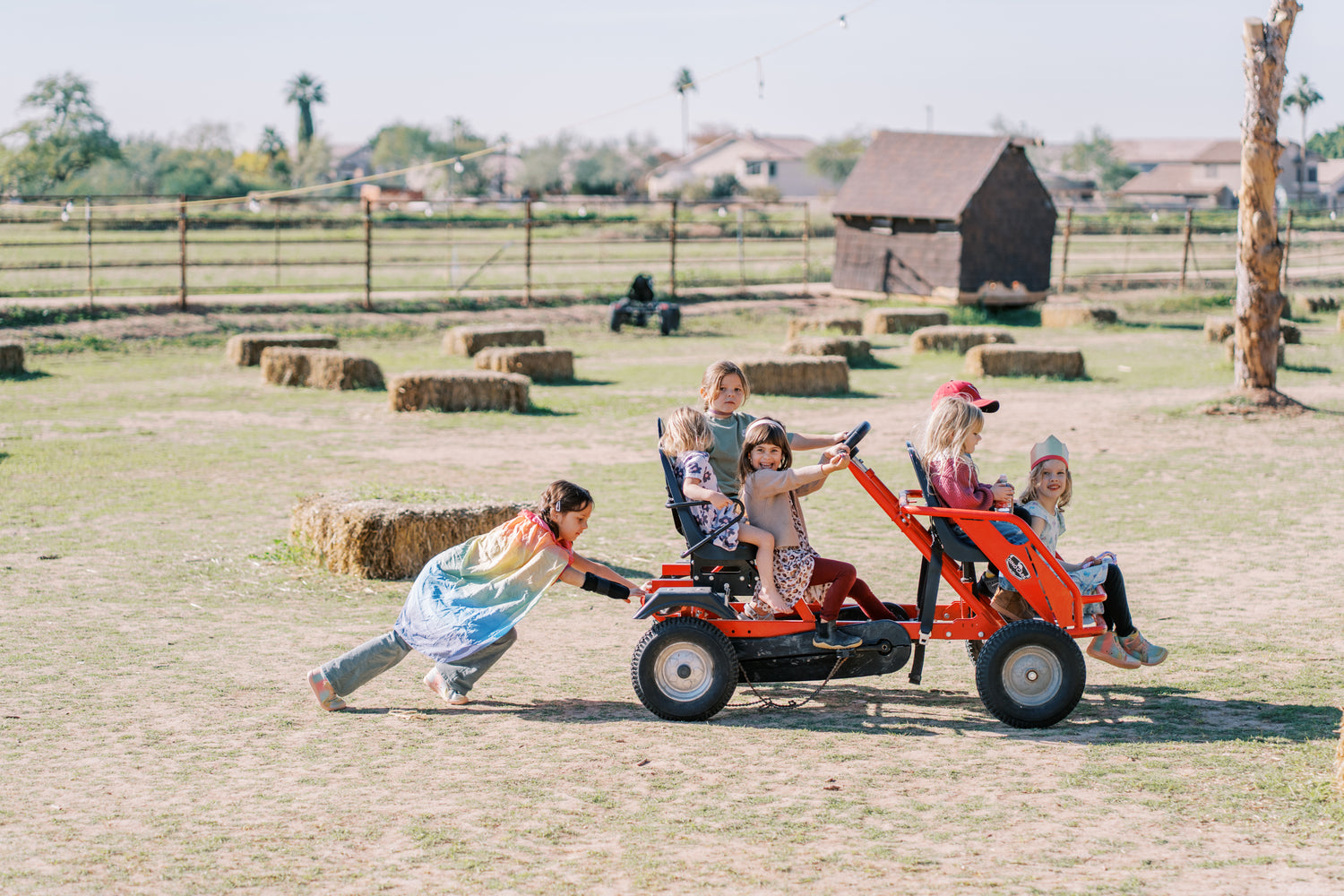 Children playing with a small tractor in an open field with hay bales and barn in the background.