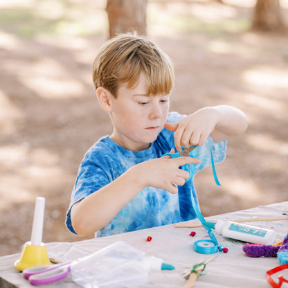 Child crafting outdoors at a wooden table with various supplies.