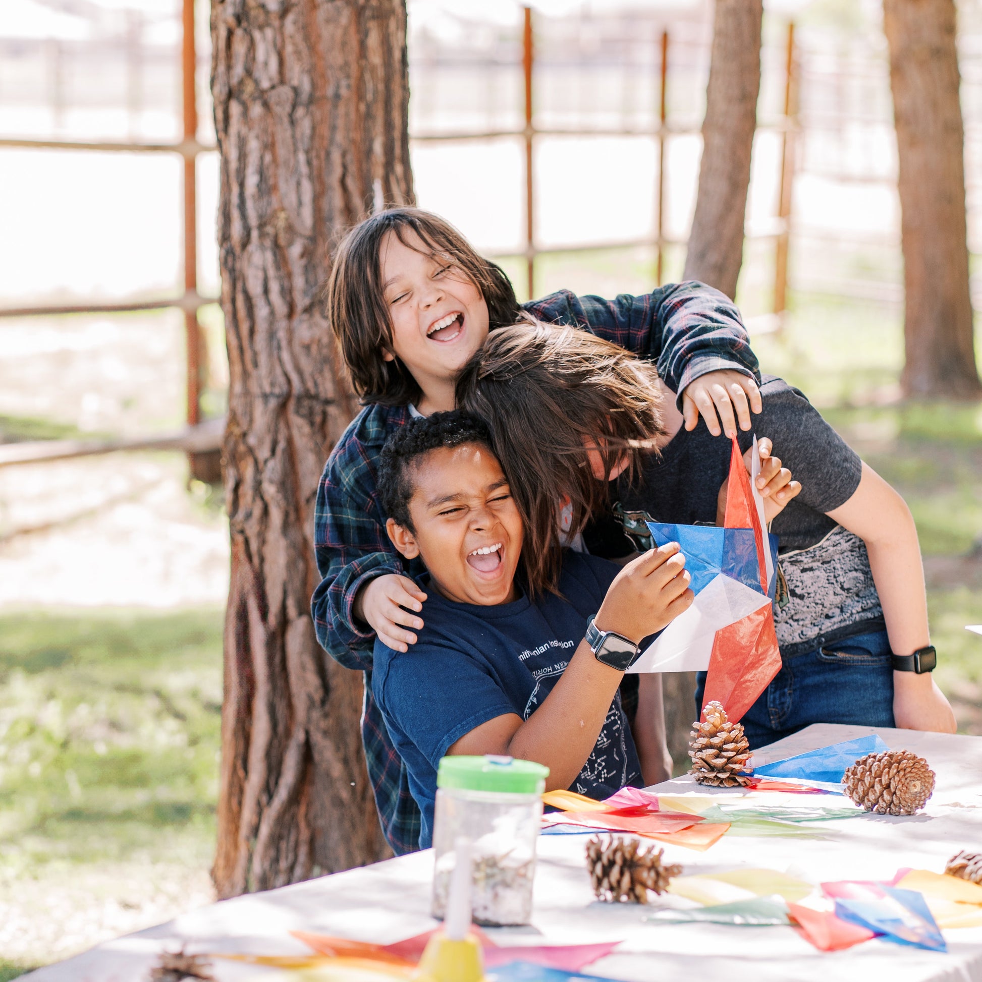 Two children playing at a table outdoors with pine cones and colorful items.