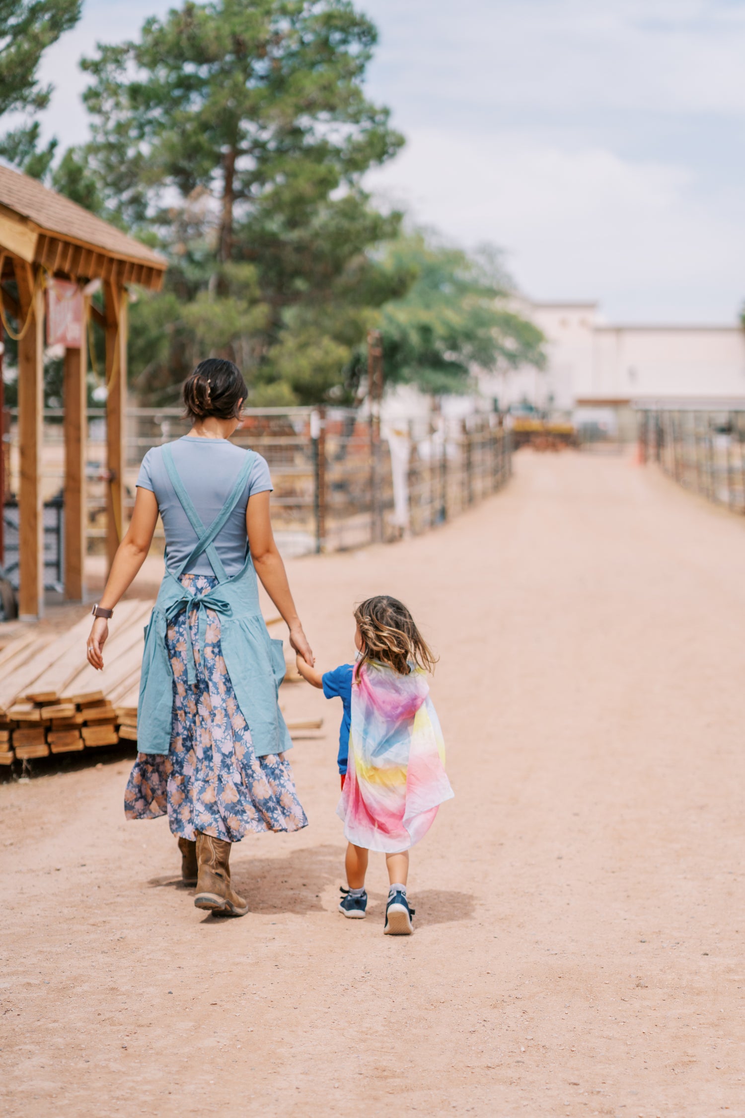 Woman and child walking hand-in-hand on a dirt path with trees and buildings in the background.