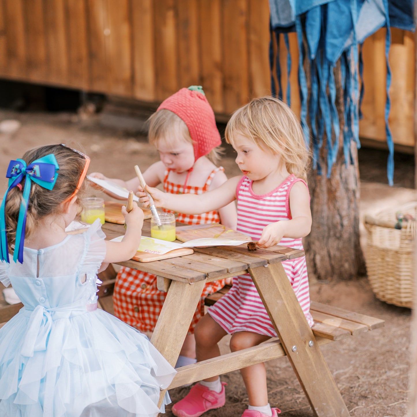 Three children in costumes sitting at a wooden picnic table outdoors.