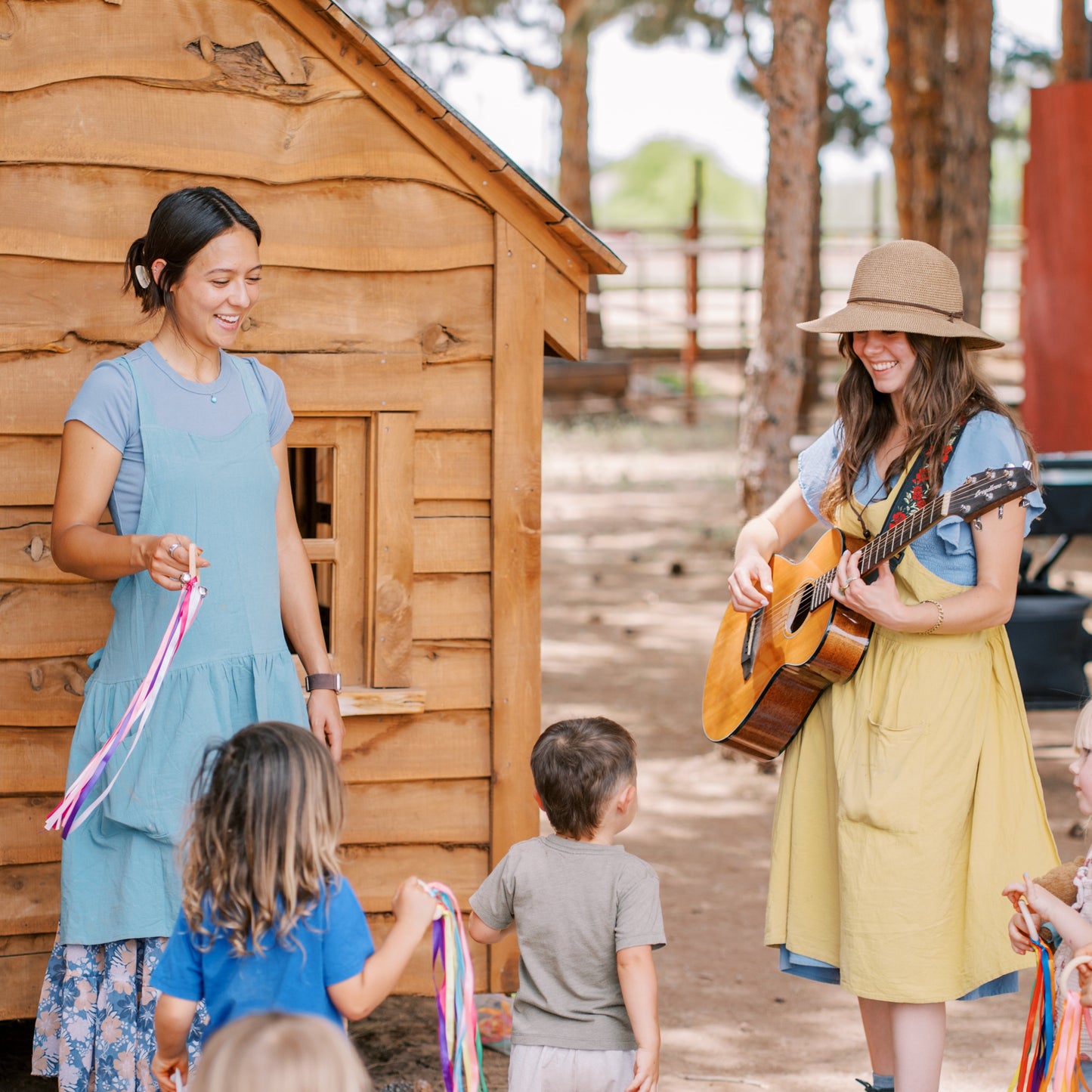 Two women with musical instruments interacting with children in front of a wooden cabin.