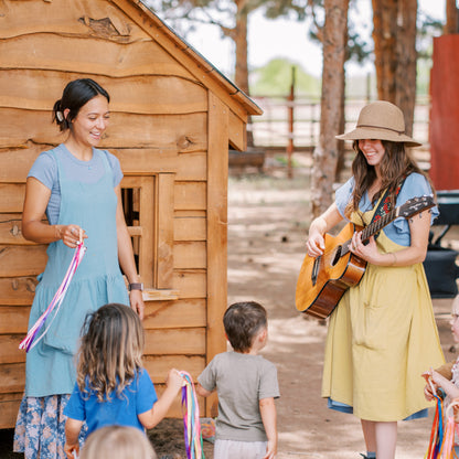 Two women with musical instruments interacting with children in front of a wooden cabin.