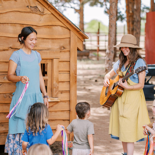 Two women with musical instruments interacting with children in front of a wooden cabin.