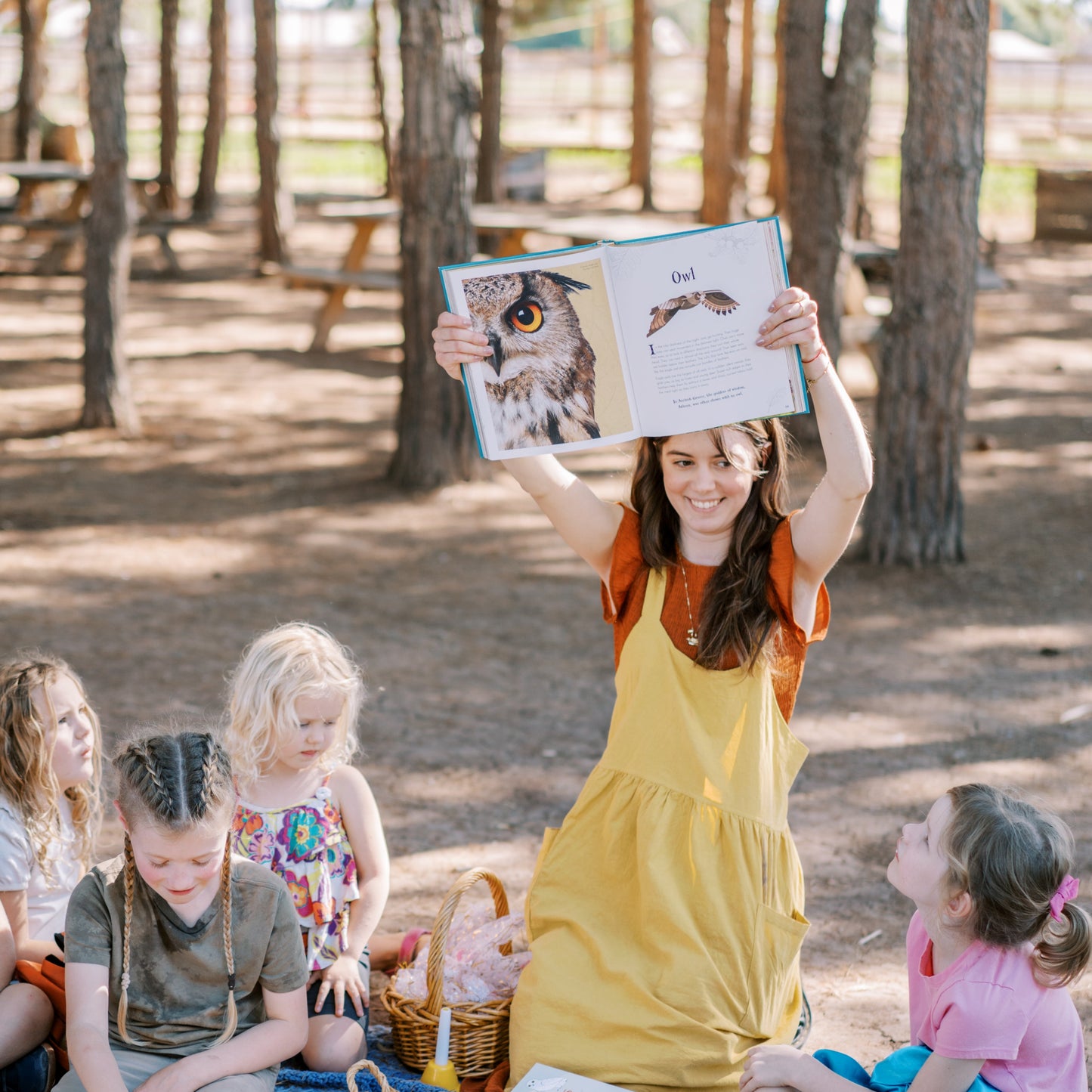 Woman reading a book to a group of children outdoors in a park.