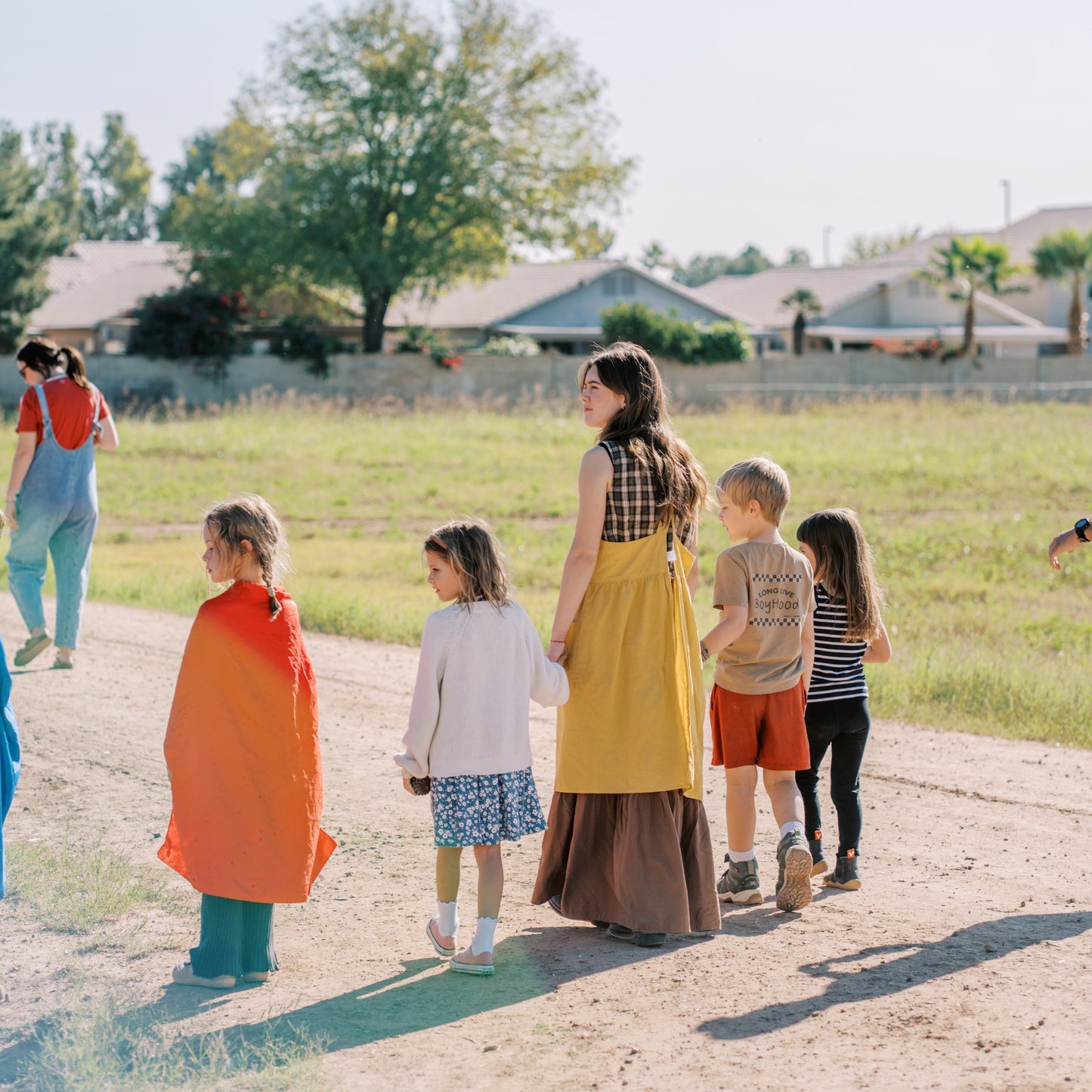 Group of people, including children, standing on a dirt path with houses and trees in the background.