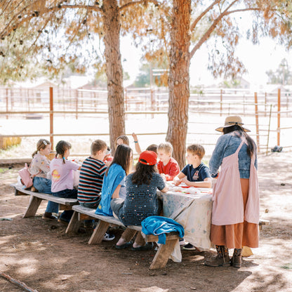 Children sitting at a table outdoors under trees with a teacher.