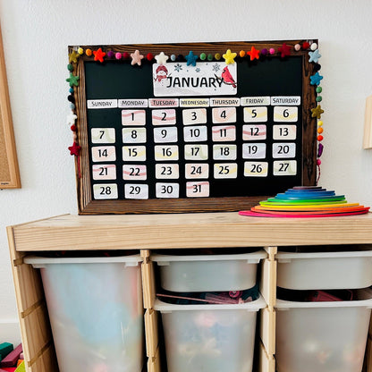 A wooden children's storage organizer with six translucent plastic bins, styled in a bright playroom. A black monthly calendar hangs on the top shelf, and colorful stacking rings and blocks are placed on and around the organizer.