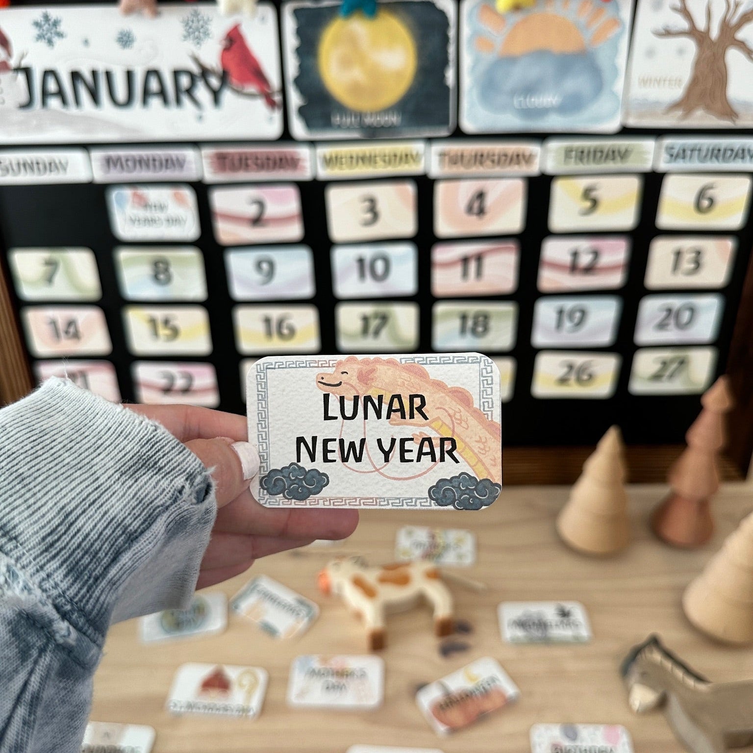 A hand holding a square card that reads 'LUNAR NEW YEAR' in front of a wooden calendar board with colorful daily tiles and educational toys on a tabletop.