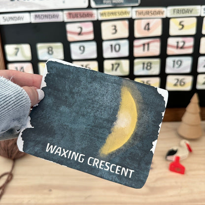 A hand holds a rustic, handmade tarot card featuring a waxing crescent moon on a dark textured background with the text 'WAXING CRESCENT'. The background shows a wooden table, a small wooden cone, and a calendar board with monthly and weekly trackers.