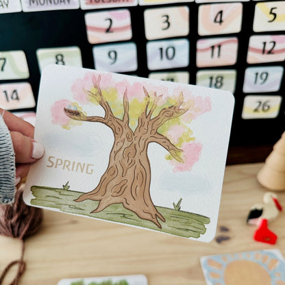 A hand holds a square card featuring a hand-drawn spring tree with pink and yellow leaves and the word 'SPRING' on a wooden tabletop. Other matching cards are visible in the background, along with a calendar and small decorative objects.